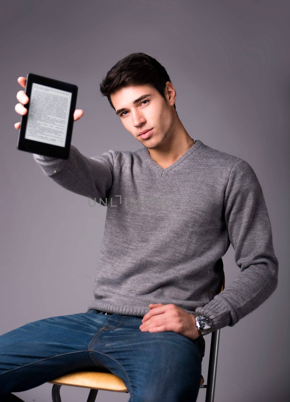 Handsome young man holding and showing ebook reader and looking at camera, sitting on grey background
