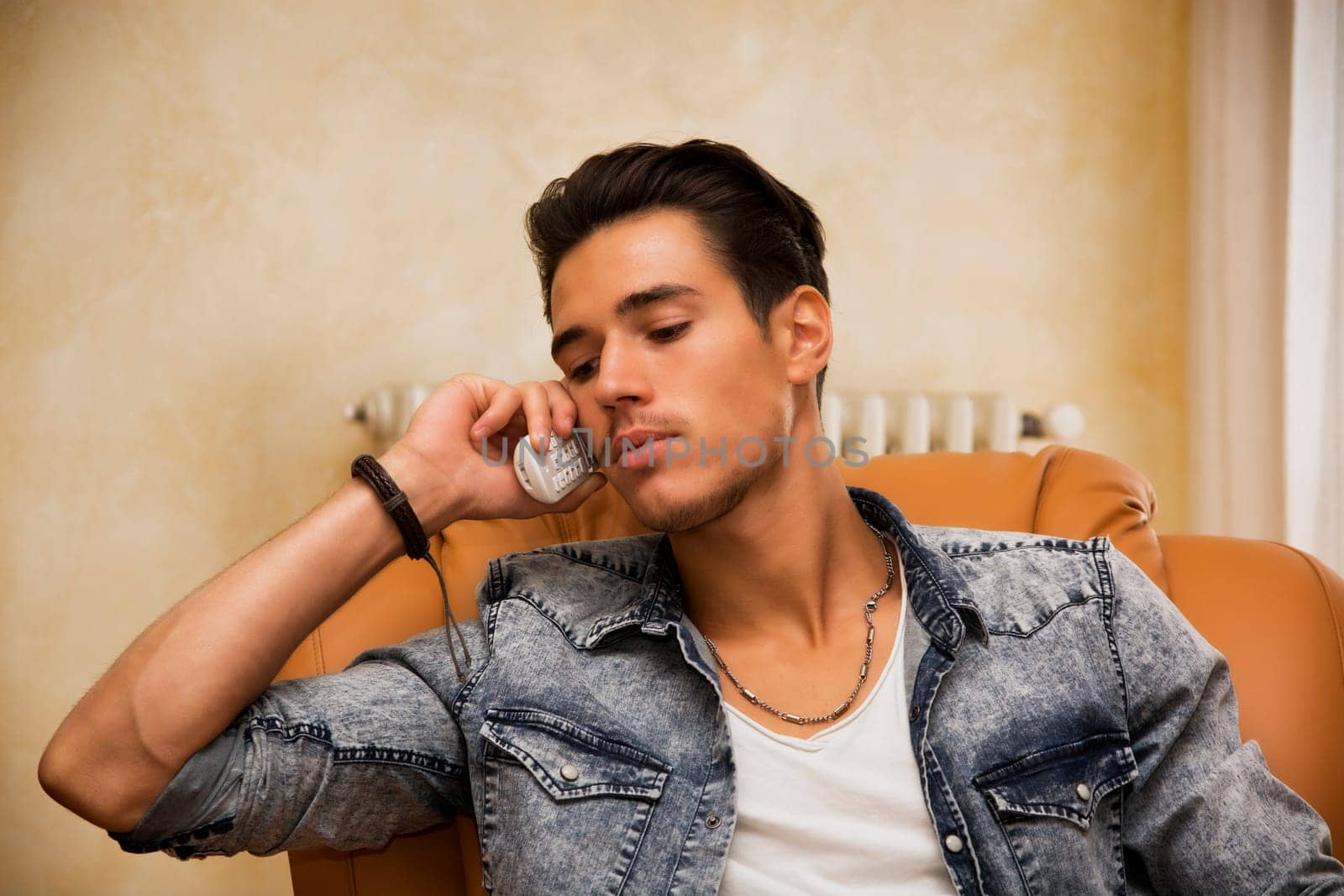 Handsome young man talking on telephone at home sitting on armchair