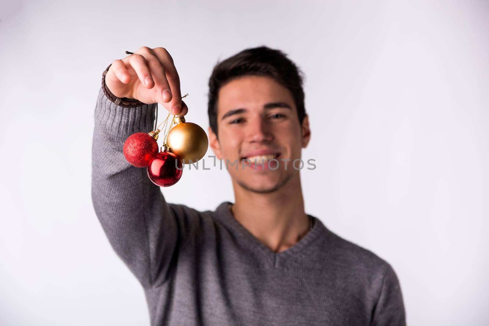 Handsome young man smiling and holding Christmas tree balls in front of his face, looking at camera on white background