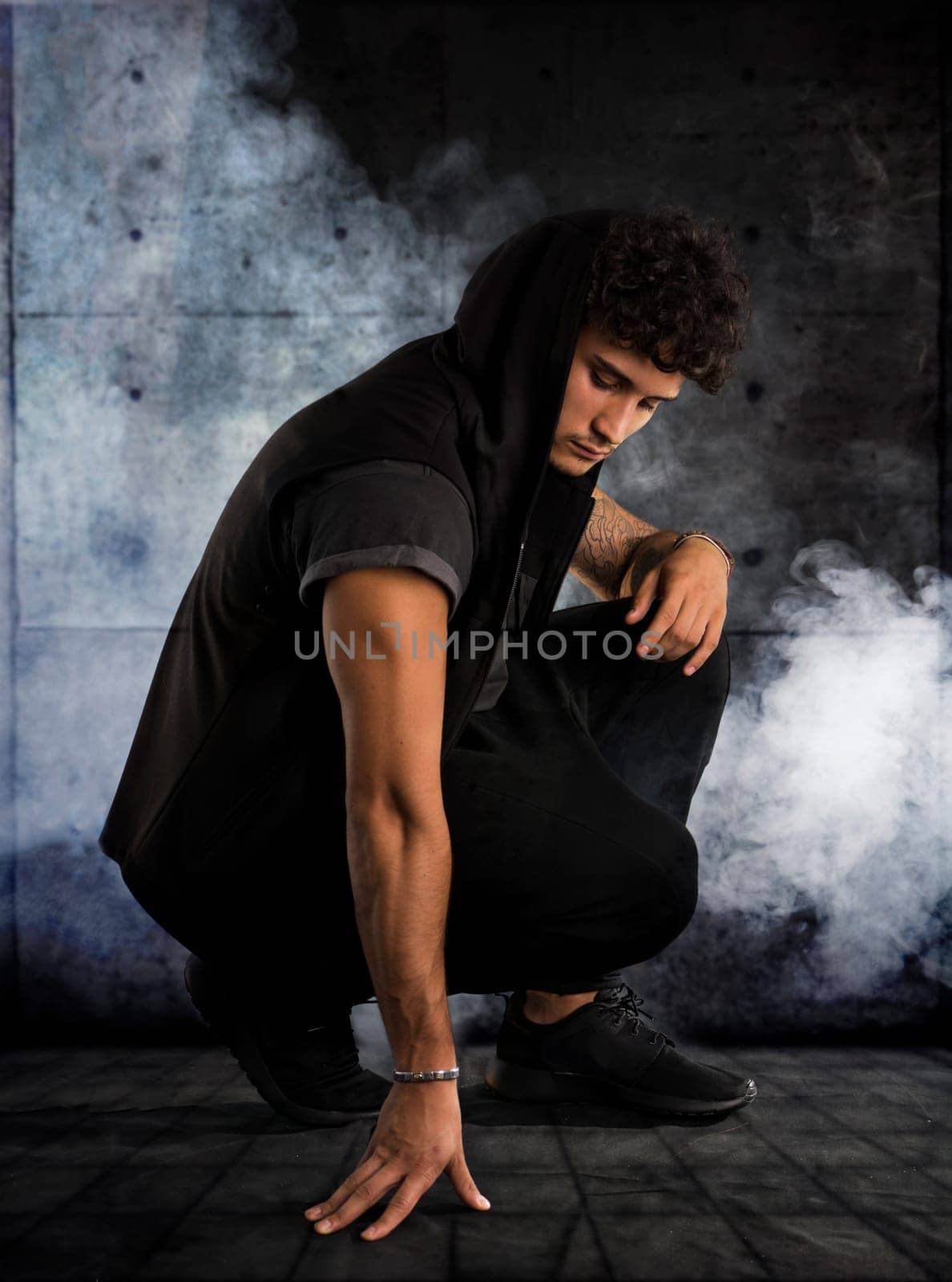 Handsome young man kneeling down in dark hoodie on black background, smoke around him. Looking down