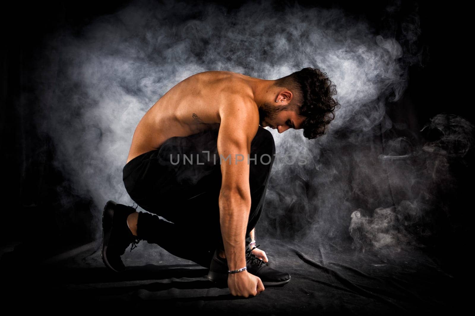 Handsome shirtless muscular young man kneeling down on black background, smoke around him. Looking down