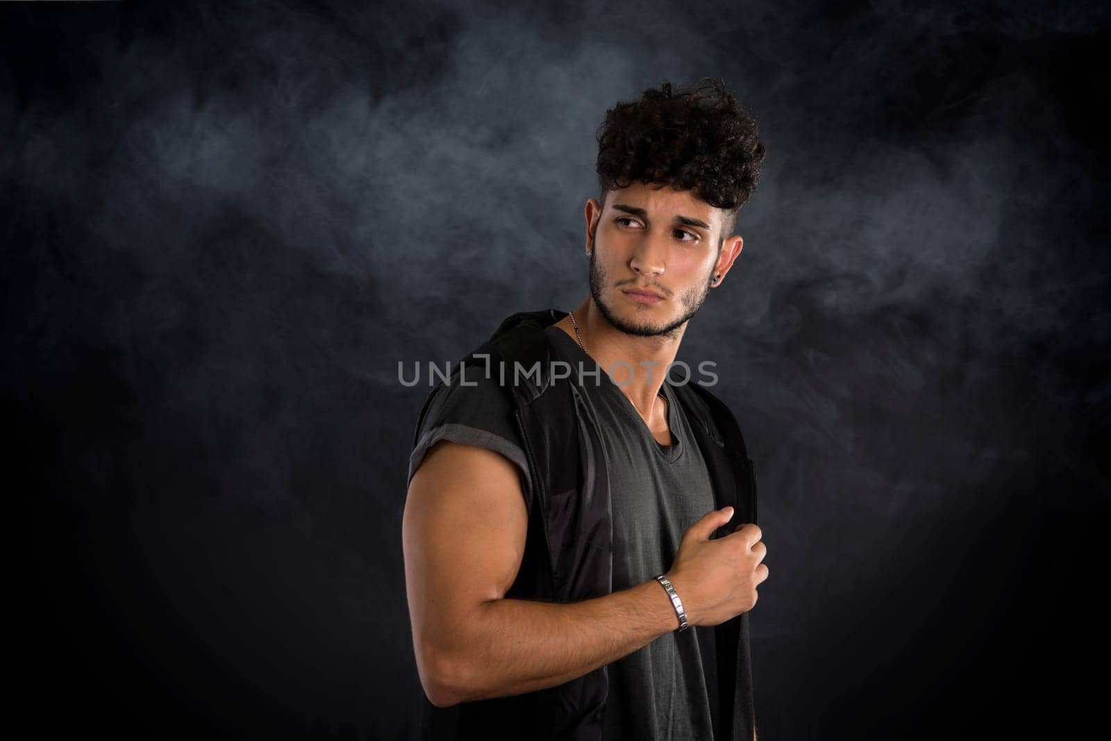 Portrait of handsome young man in dark t-shirt on black background, with smoke around. Looking away