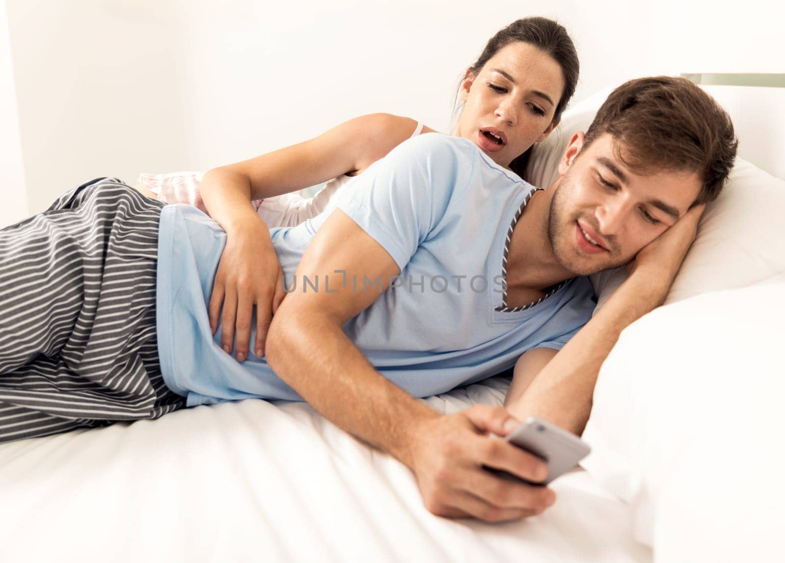 Addicted young man on bed texting while woman looks angry