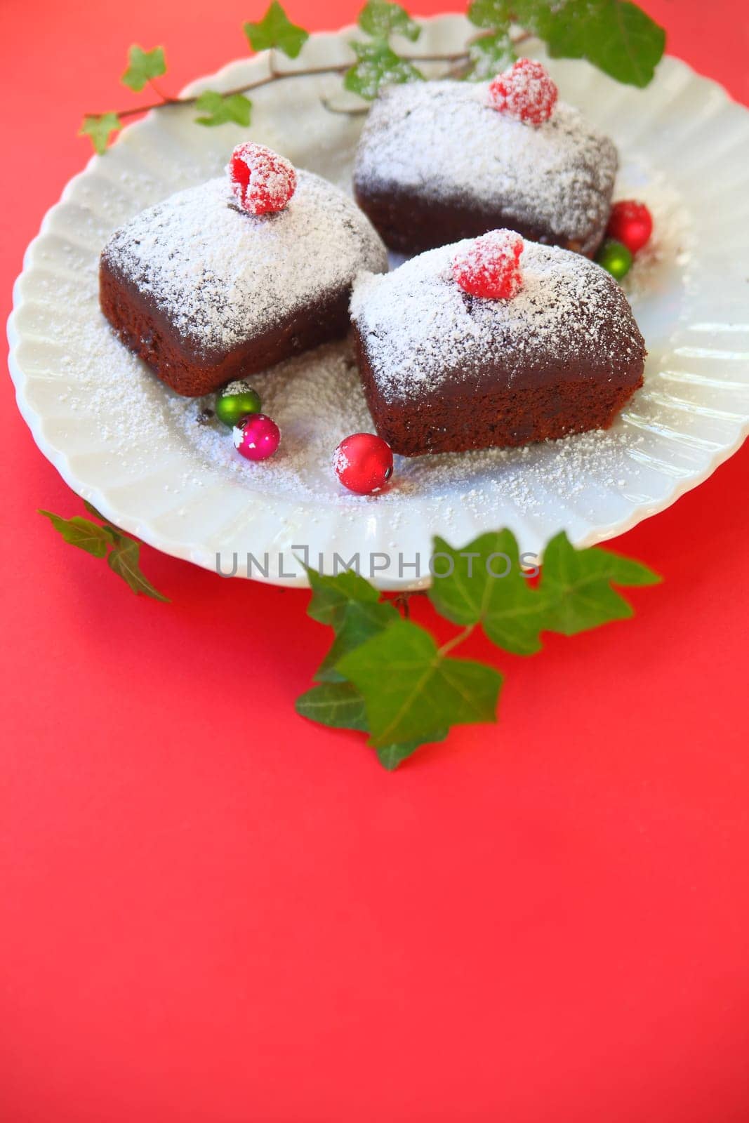 Vertical view of mini chocolate cakes dusted with icing or powdered sugar, topped with fresh raspberries on a plate with tiny ornaments and ivy leaves
