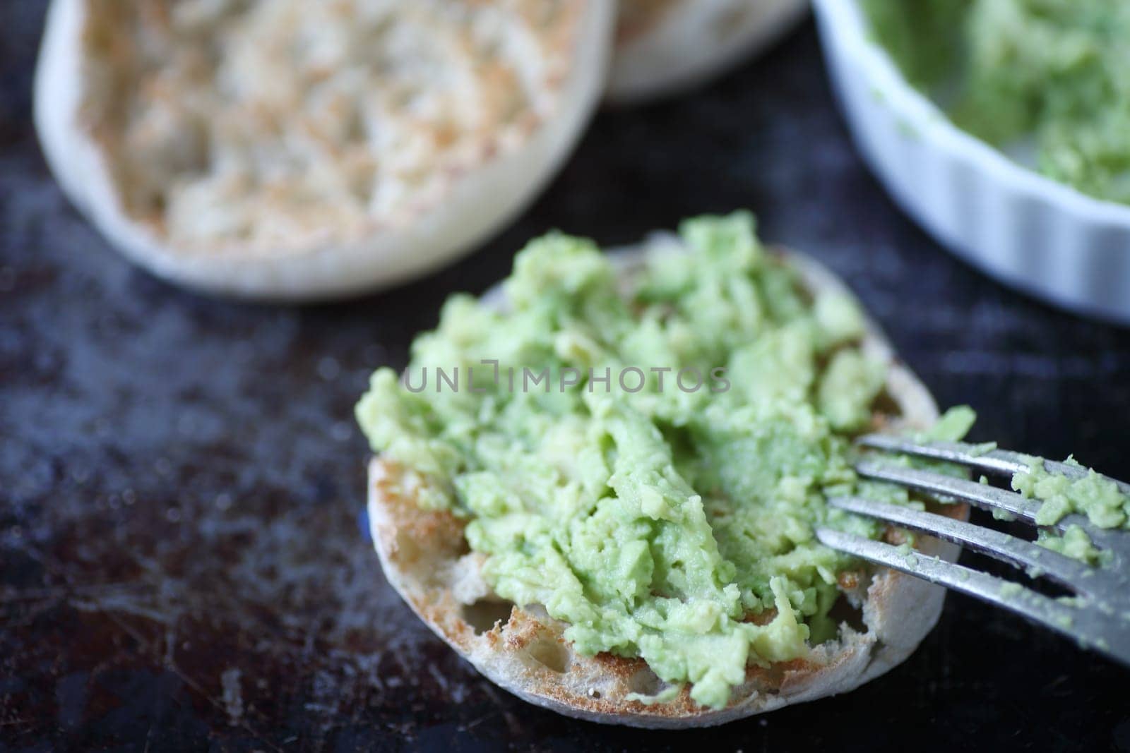 Toasted English muffins with mashed avocado on metal surface