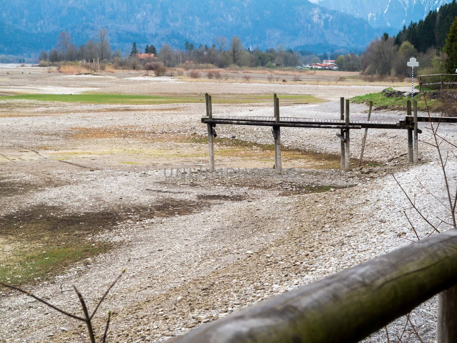 View of the drained Forggensee with Schwangau behind it near Füssen / Bavaria with wooden walkway in the foreground. The Forggensee is fed by thawing snow from the high Alps. The sedimentary rock carried with it settles at the bottom of the lake. In order to remove this sedimentary rock,the water of the lake is drained to make it possible to transport it with construction vehicles. By the summer,the lake has returned to its normal water level and is used by excursion boats and recreational boats.