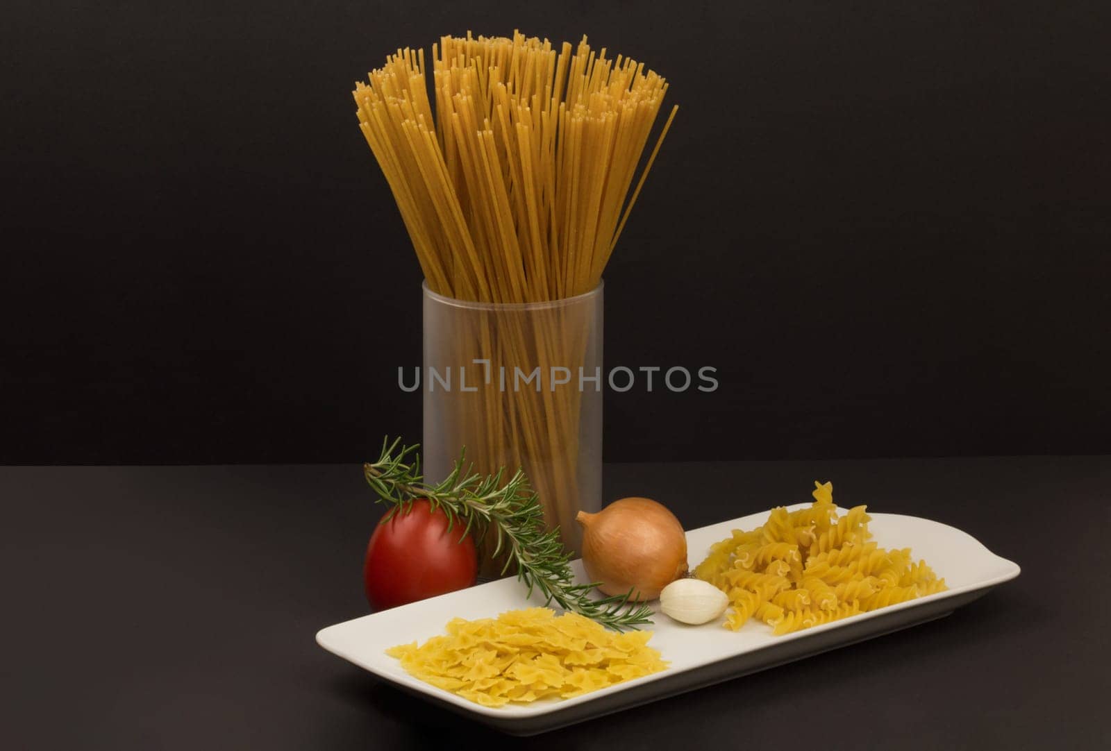Glass with raw spaghetti and other pasta variants and vegetables on a plate.