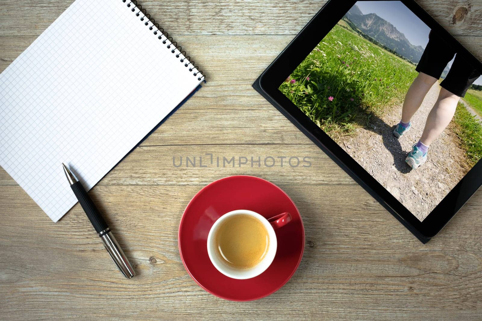 Tablet with legs of a woman on a hike in the mountains with meadow and block with ballpoint pen