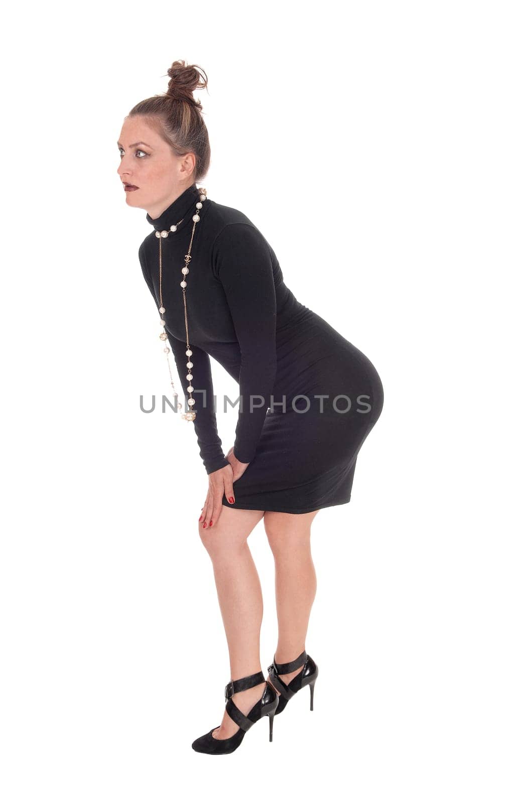 A beautiful young woman in a black dress and a necklace standing in.profile, bending forwards, isolated for white background..