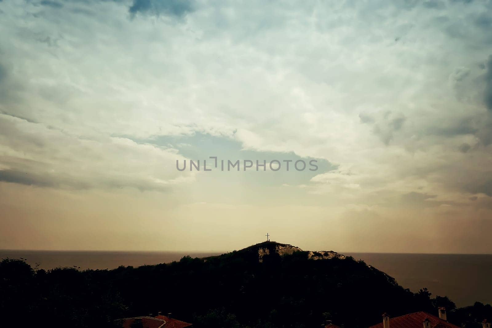 A silhouette of a holy christian cross illuminated by sunlight on top of a hill on a dramatic cloudy sky. Beautiful view at the coast of the Black Sea in Balchik city, Bulgaria.