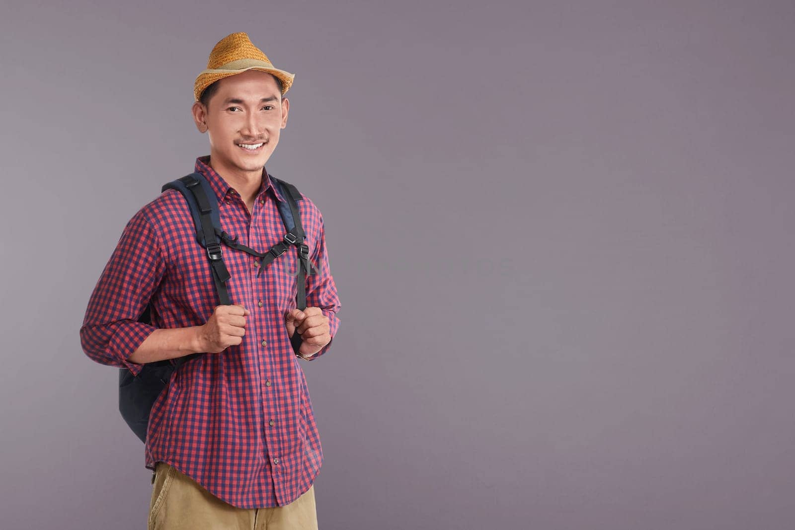 Handsome asian tourist with hat carrying backpack against dark background
