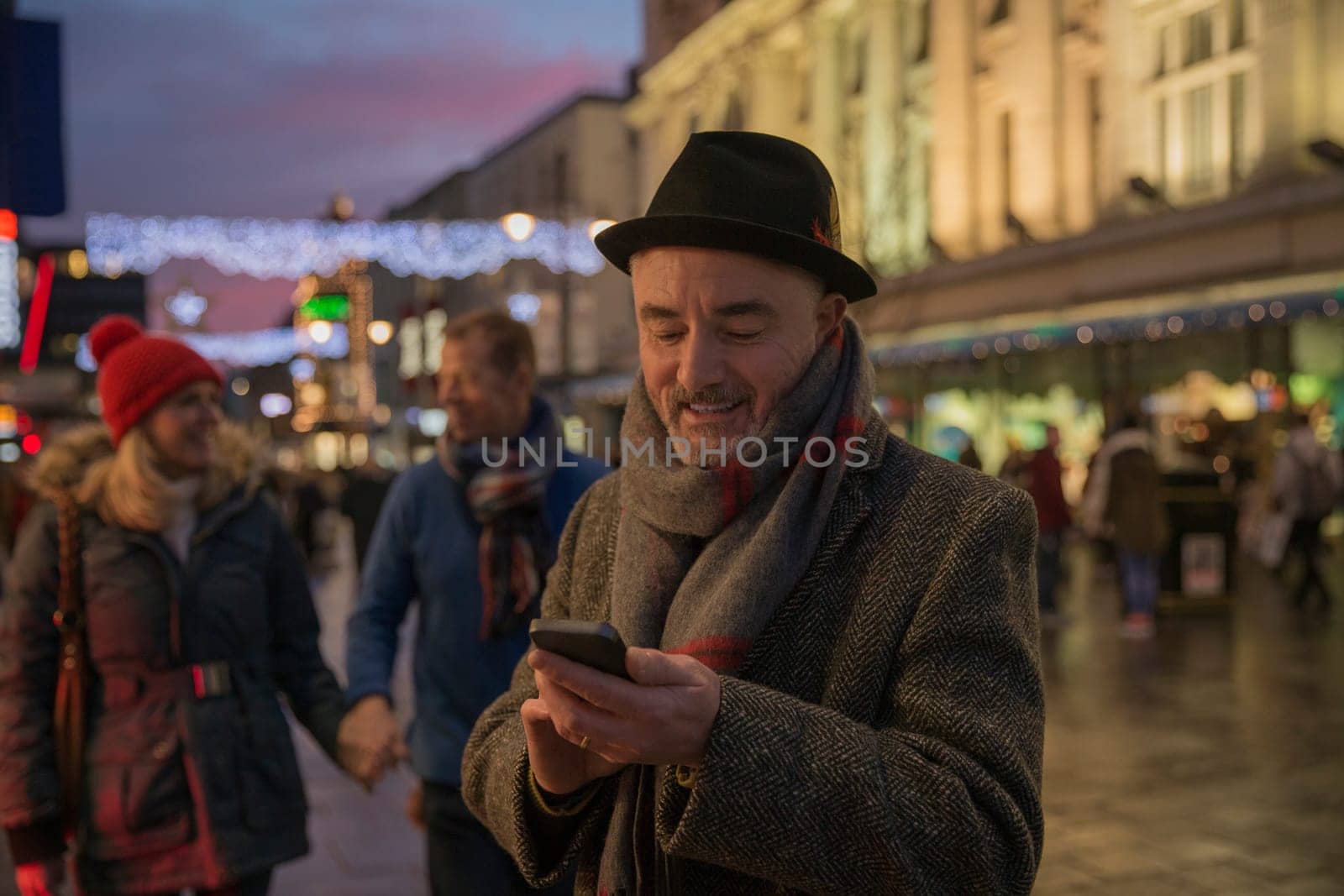 Mature man is enjoying an evening stroll through town at Christmas time. He has stopped to use his smart phone.