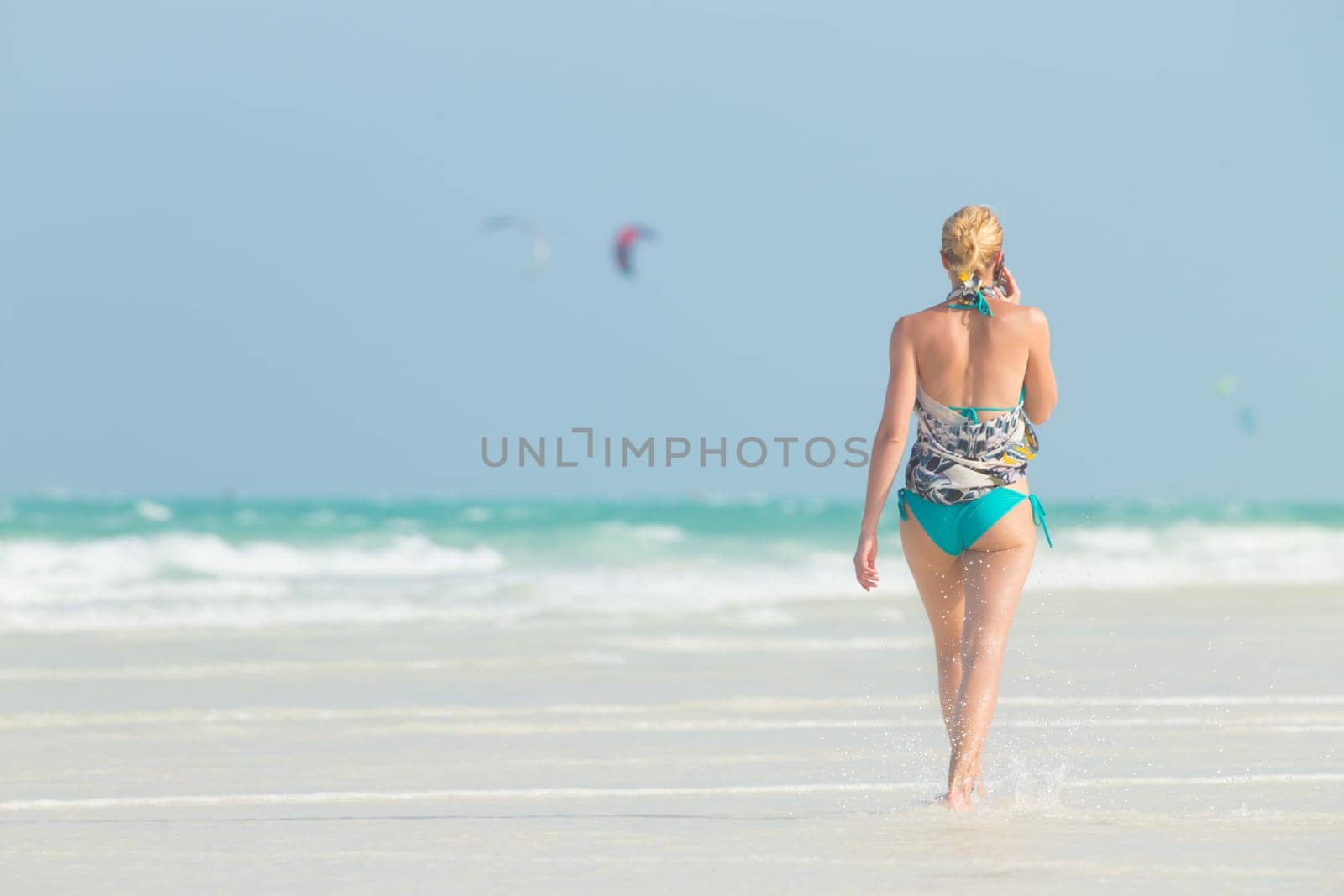 Happy woman having fun, enjoying summer, walking joyfully on tropical beach. Beautiful caucasian model wearing colorful scarf and turquoise bikini on vacations on Paje beach, Zanzibar, Tanzania