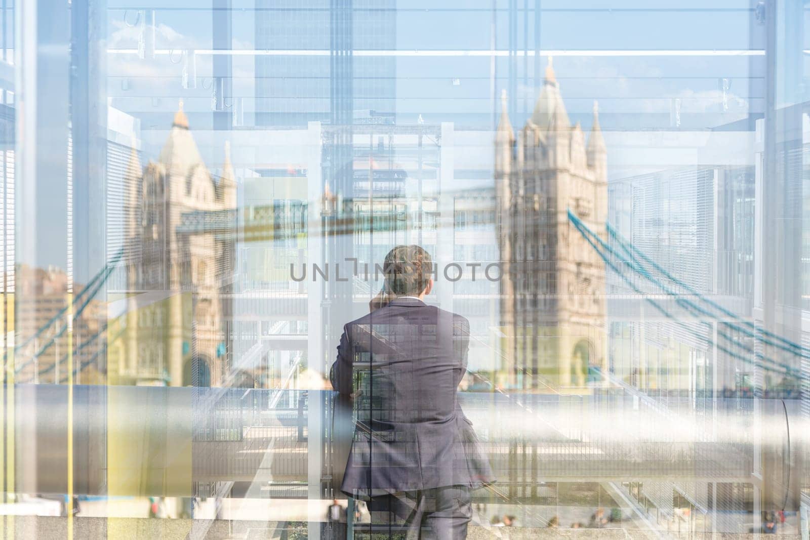 Businessman talking on mobile phone while looking trought the business office window at Tower Bridge in London city, UK.
