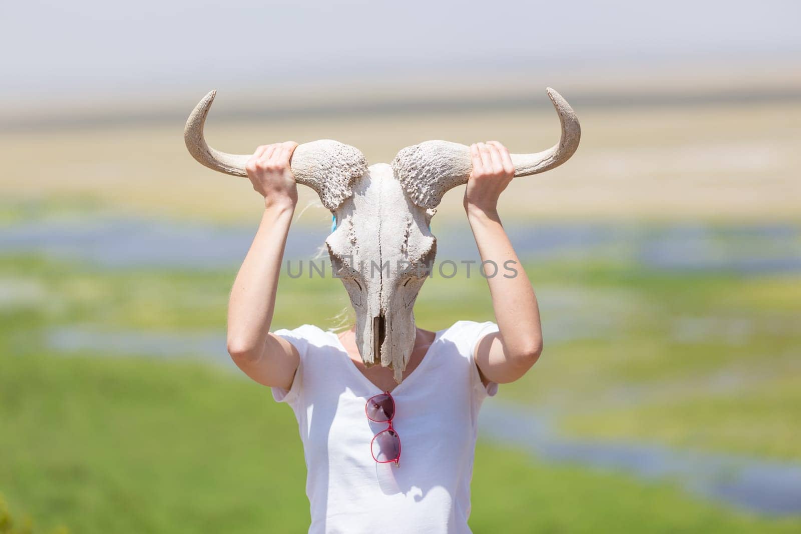 Woman holding a white wildebeest skull wearing it like a mask in nature on african wildlife safari, Amboseli national park, Kenya.