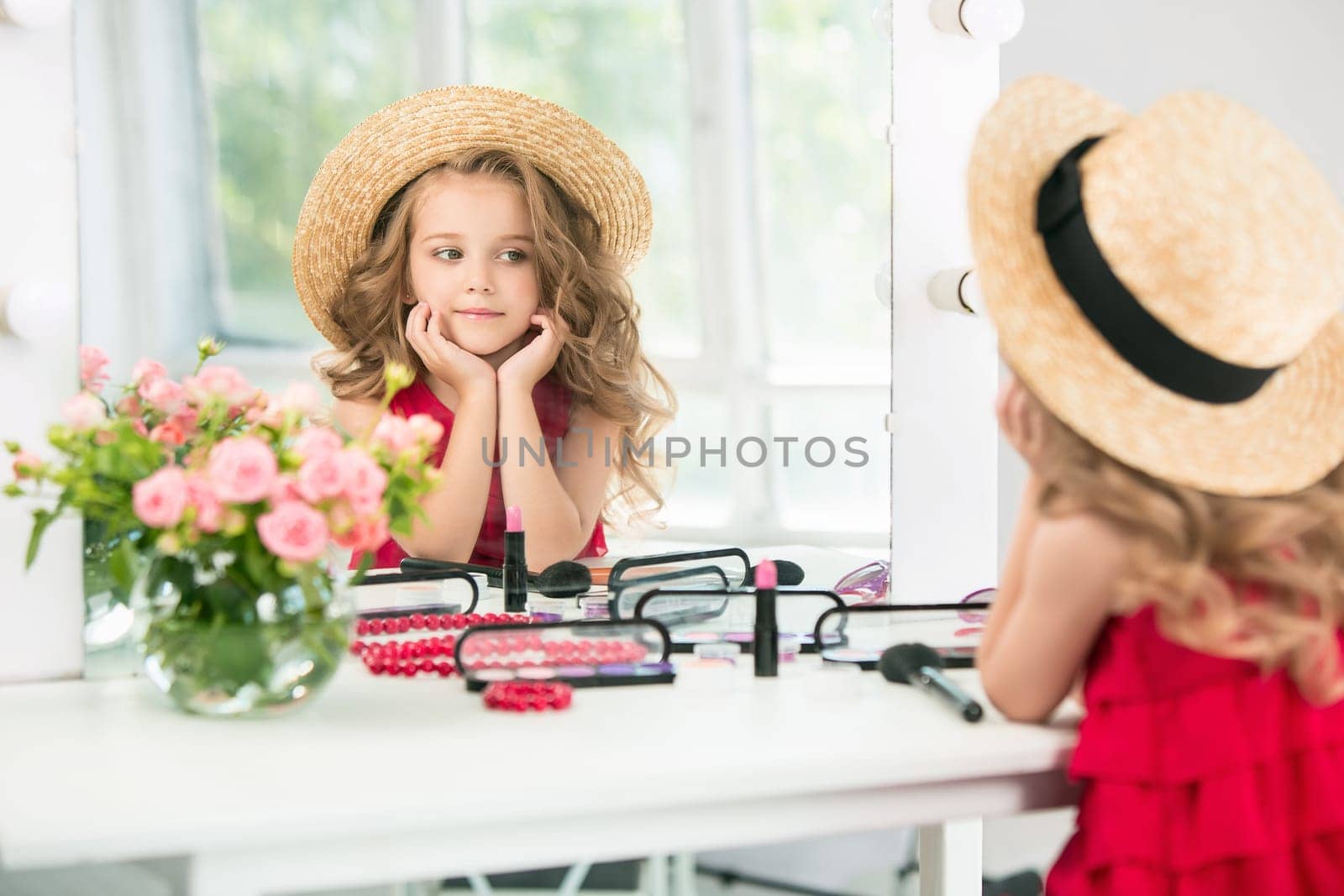 A little girl with red dress and cosmetics. She is in mother's bedroom, sitting near the mirror.