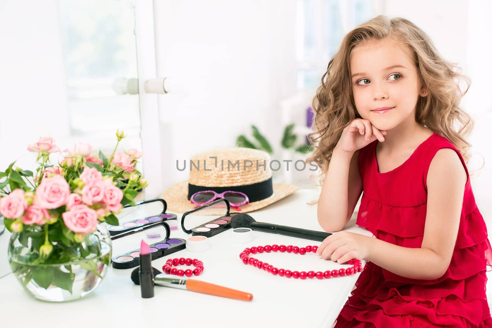 A little girl with red dress and cosmetics. She is in mother's bedroom, sitting near the mirror.