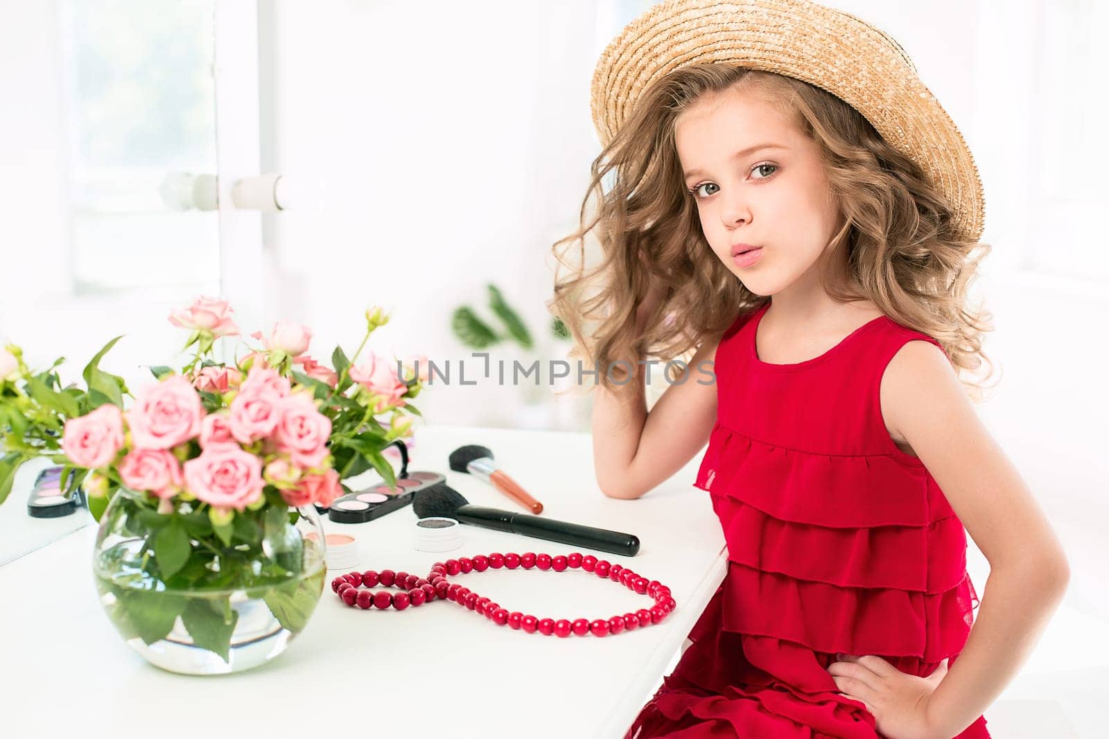 A little girl with red dress and cosmetics. She is in mother's bedroom, sitting near the mirror.