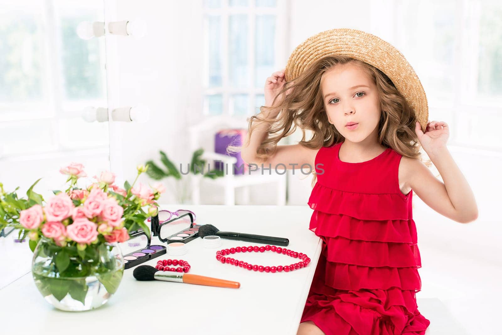 A little girl with red dress and cosmetics. She is in mother's bedroom, sitting near the mirror.
