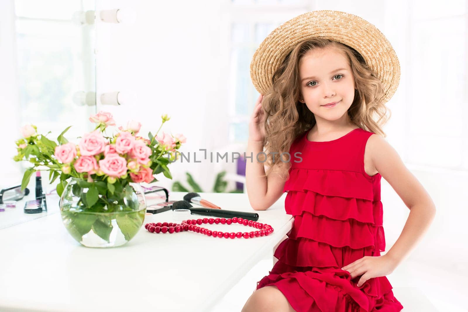 A little girl with red dress and cosmetics. She is in mother's bedroom, sitting near the mirror.