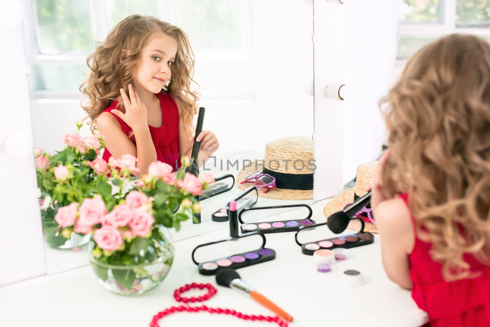 A little girl with red dress and cosmetics. She is in mother's bedroom, sitting near the mirror.