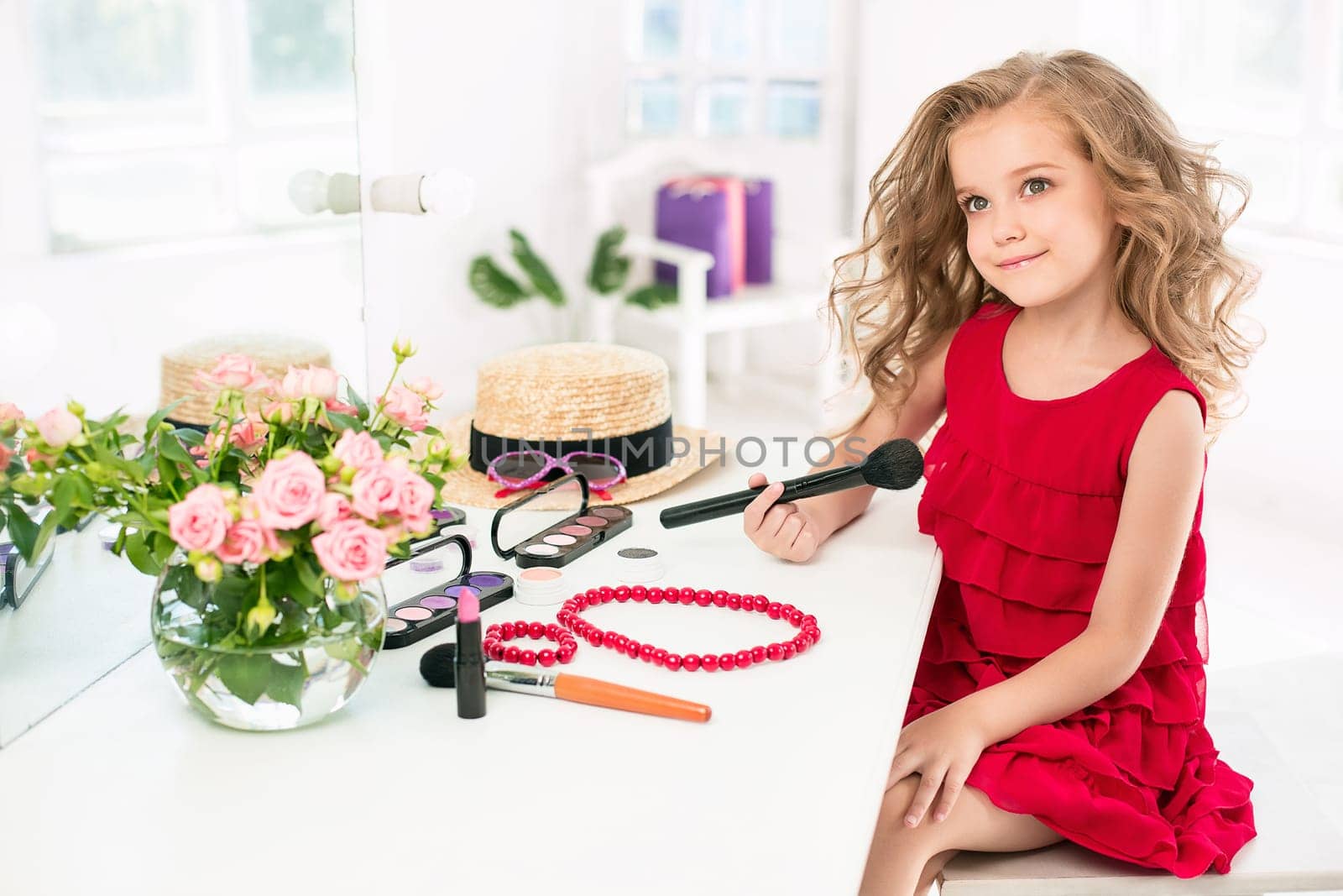 A little girl with red dress and cosmetics. She is in mother's bedroom, sitting near the mirror.