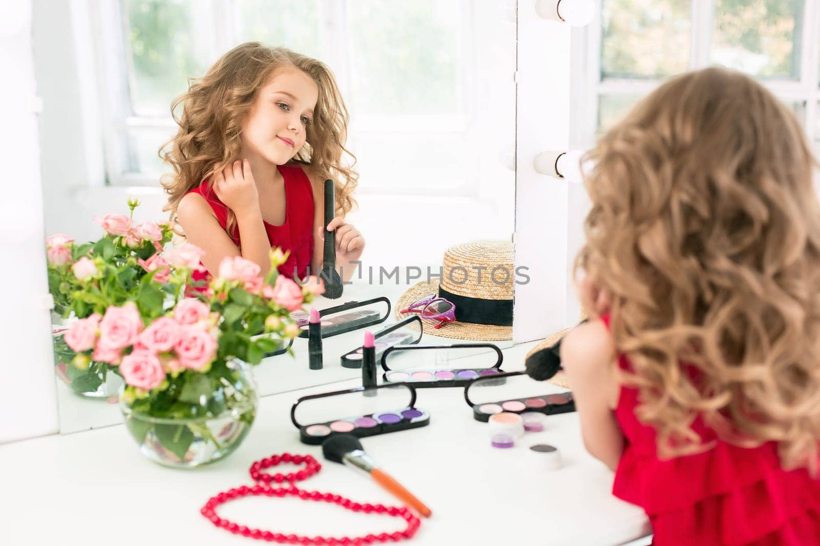 A little girl with red dress and cosmetics. She is in mother's bedroom, sitting near the mirror.