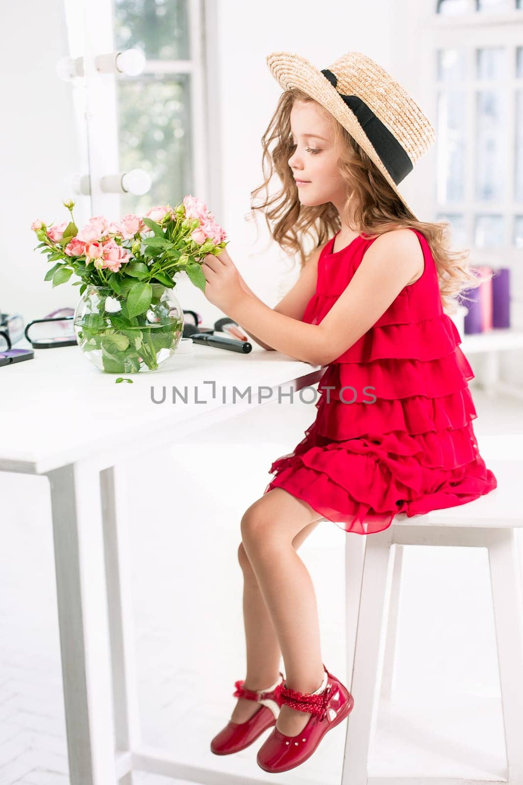 A little girl with red dress and cosmetics. She is in mother's bedroom, sitting near the mirror.