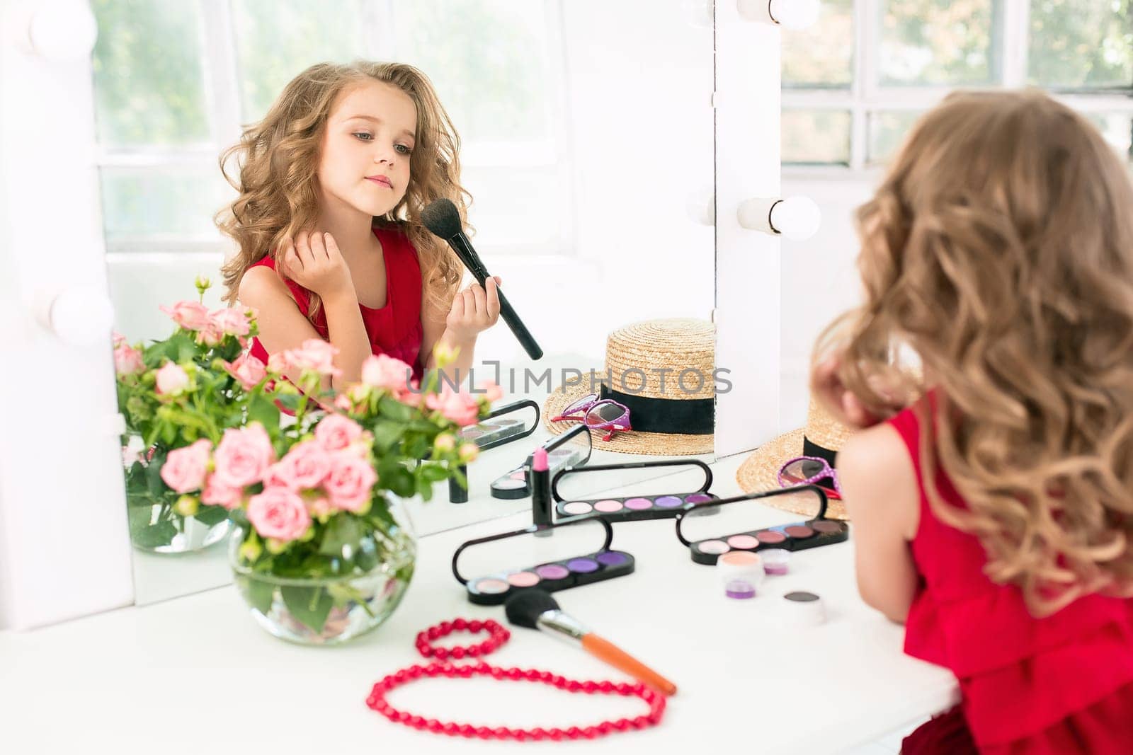 A little girl with red dress and cosmetics. She is in mother's bedroom, sitting near the mirror.