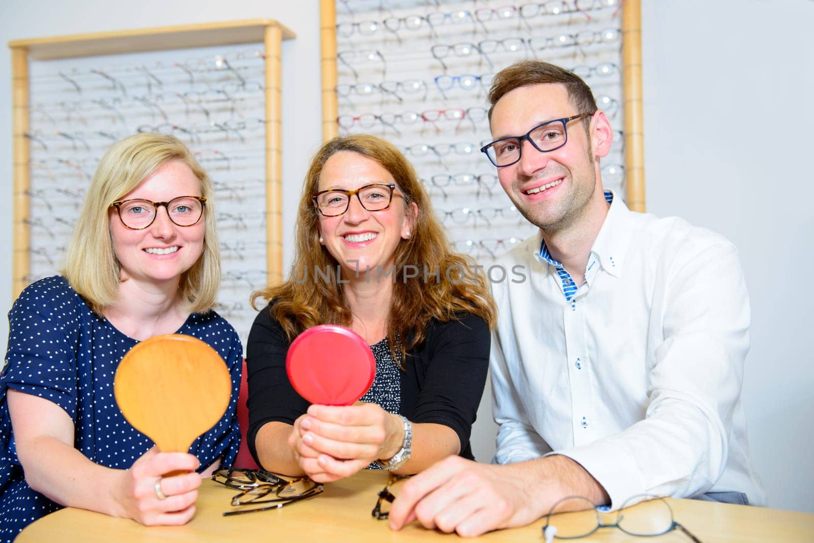 in optician shop-two women and one man with hand mirror up selecting new glasses