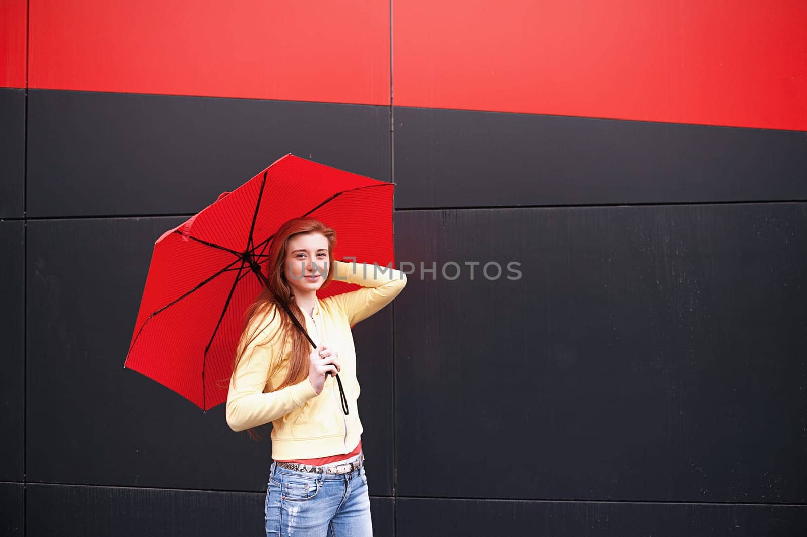 A pretty girl with long red hair, with a red umbrella in her hands, walks and smiles. Portrait in the open air