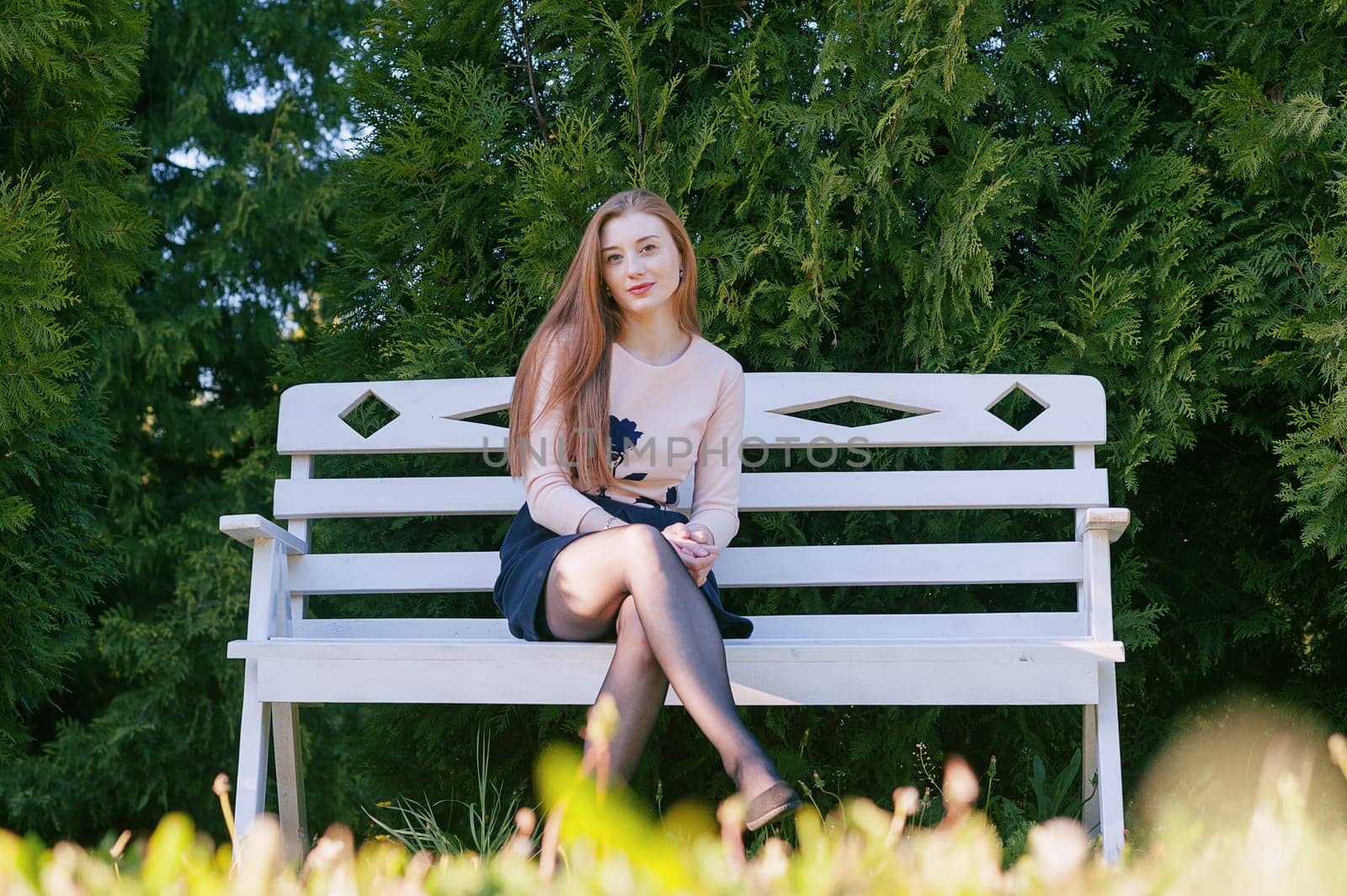 A cute girl with long red hair, sits on a white bench, against a backdrop of a garden with thujas, and smiles. Portrait in the open air