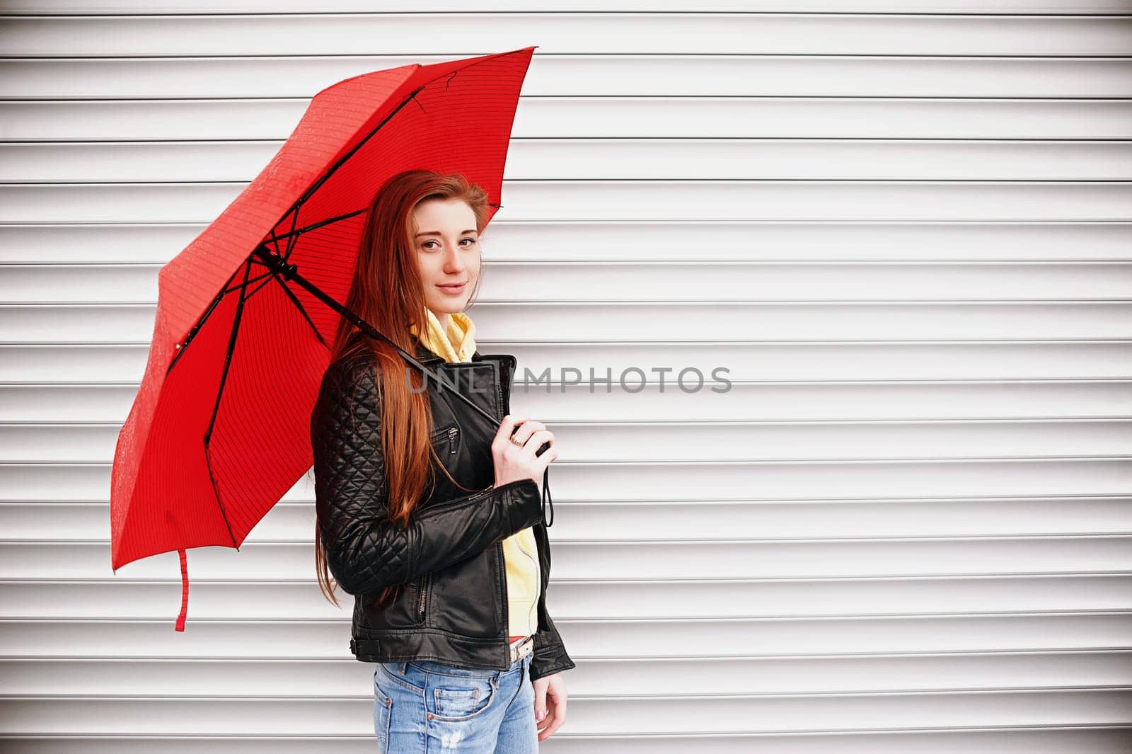 A pretty girl with long red hair, in a black leather jacket with a red umbrella in her hands, walks and smiles. Portrait in the open air