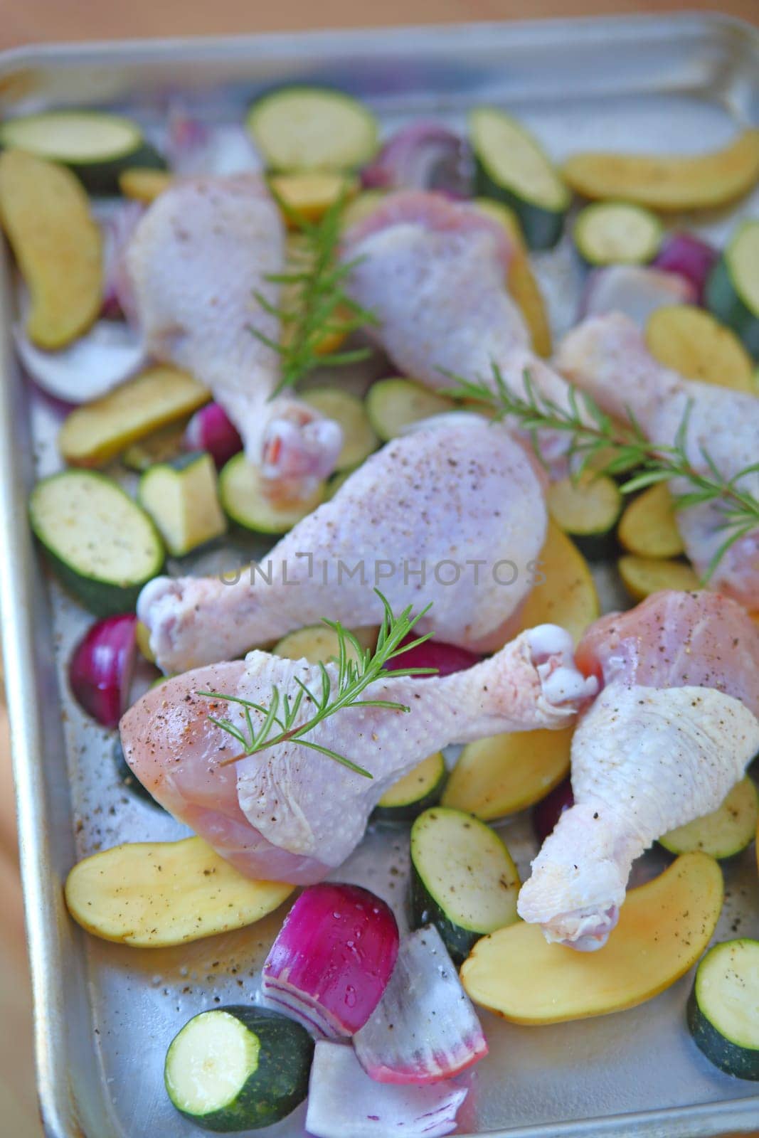 Baking sheet with chicken legs, zucchini, rosemary, onions, and potatoes before going in the oven
