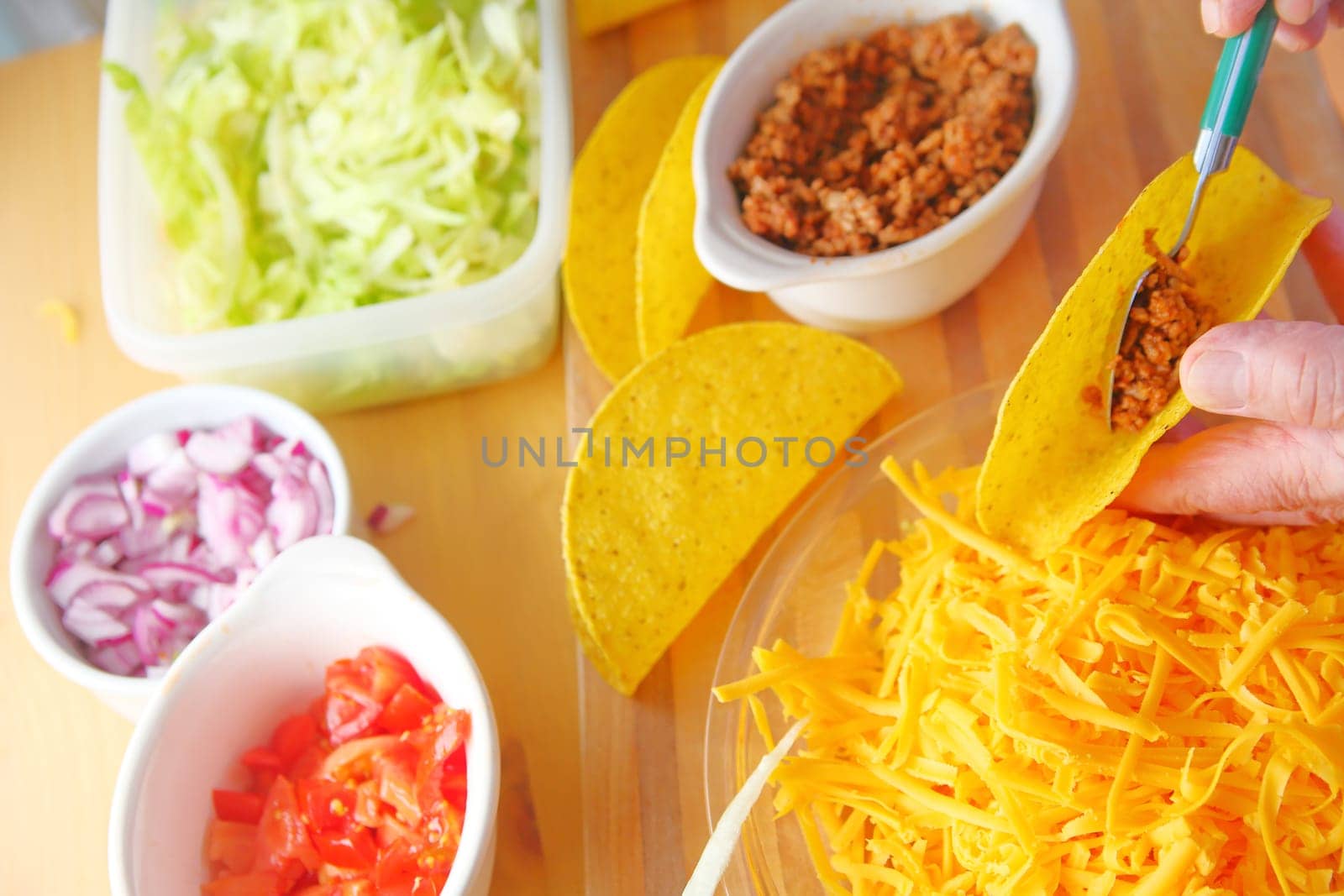 A man puts ground beef into a taco shell with grated cheese, shredded lettuce, chopped tomatoes and onions nearby