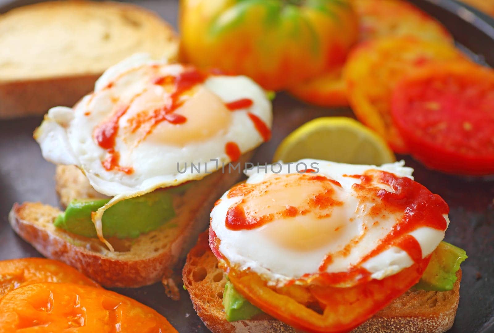 Sliced avocados on toast with fried eggs, heirloom tomatoes and hot chili sauce