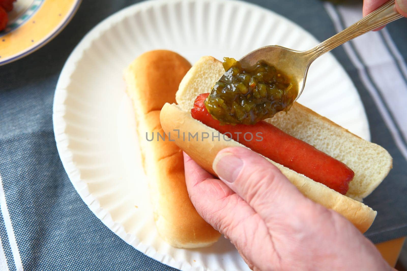 A man adds sweet pickle relish to a hot dog over a paper plate with additonal bun