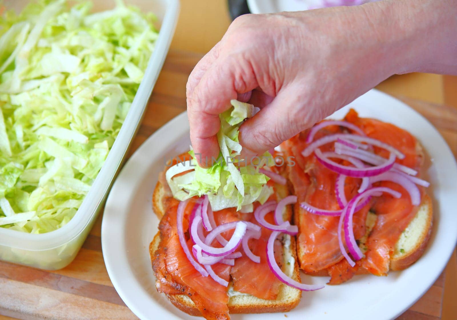 A man puts shredded lettuce on a smoked salmon sandwich