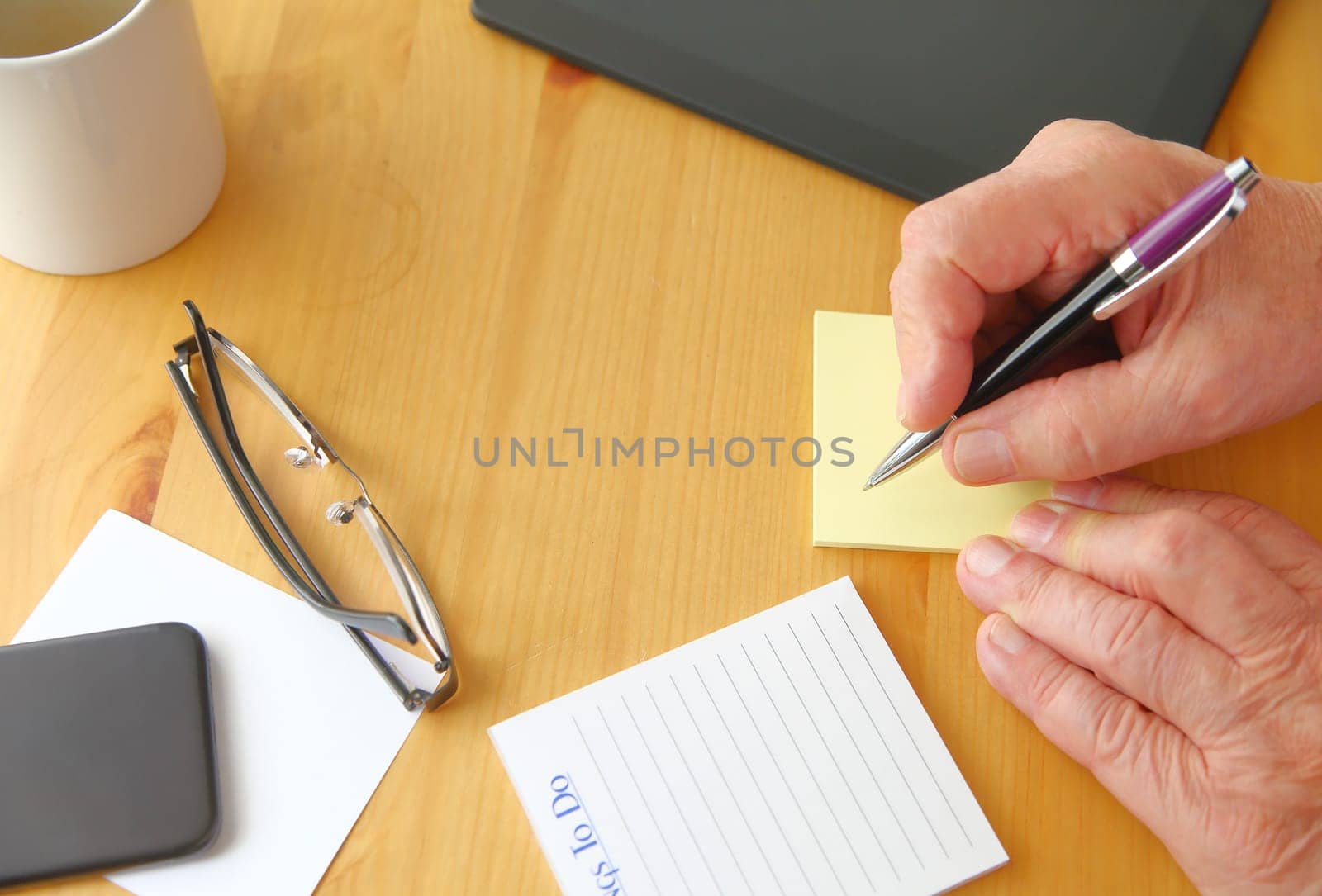 A man holds a pen over a desk with tablet, phone, notepads, glasses, coffee cup and copy space