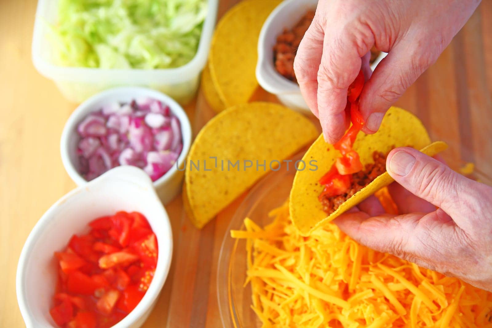 Overhead of a man making tacos with ground beef, tomatoes, onions, lettuce and grated cheese alongside