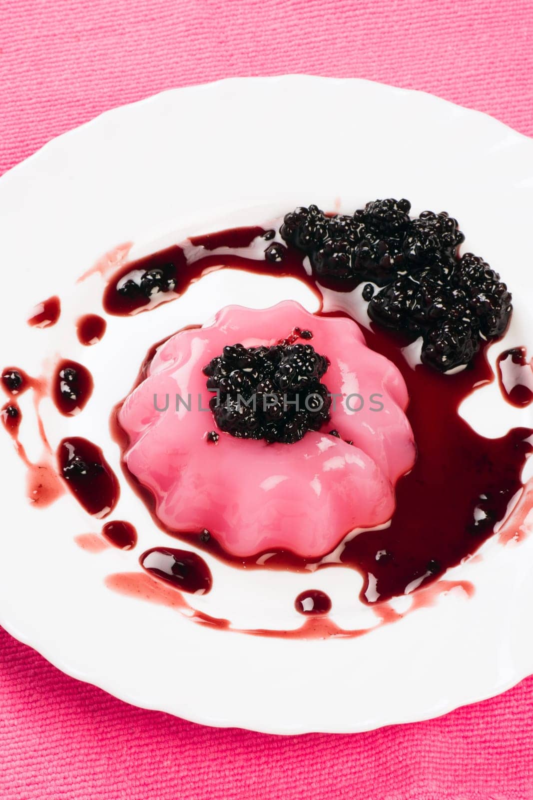 Pudding with sauce of blackberry is on white plate on pink background.