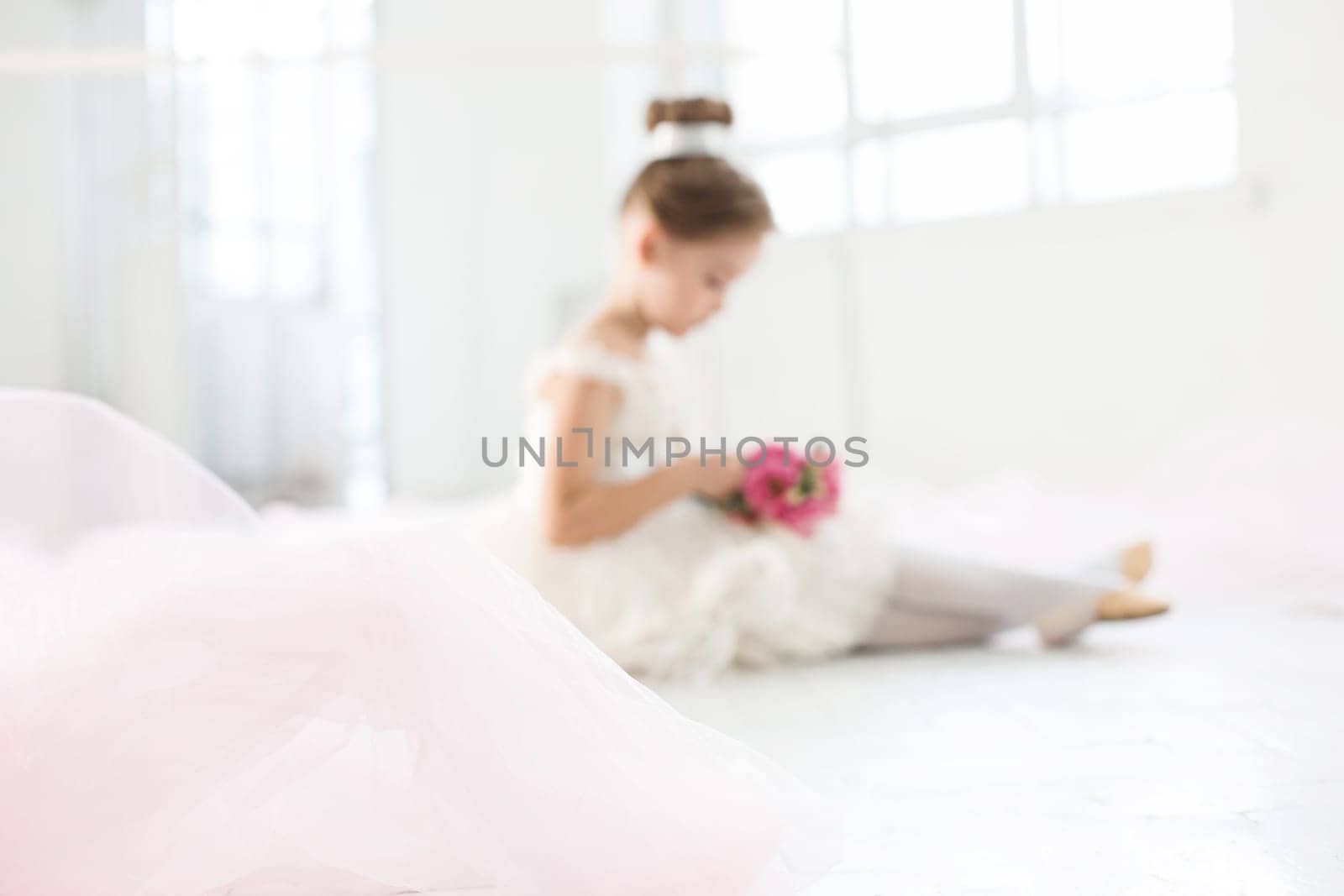 The little balerina in white tutu in class at the ballet school sitting on the wooden floor with flowers