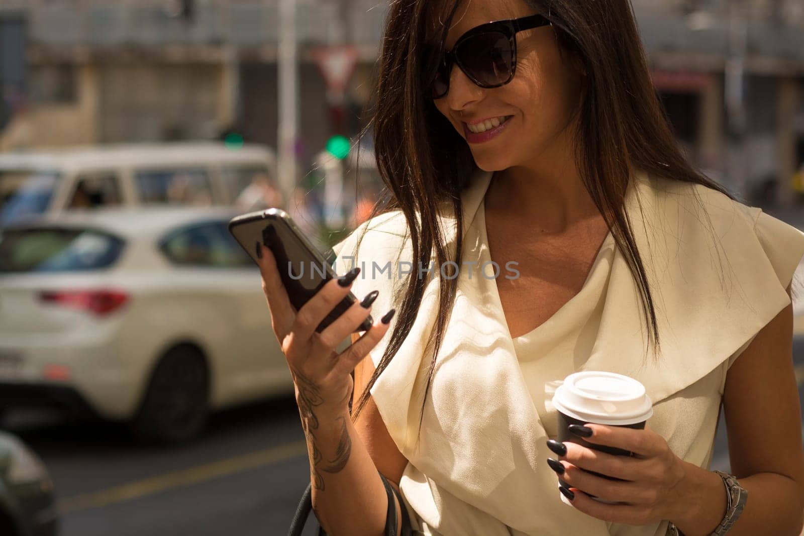 Fashionable young brunette wearing white dress and sunglasses laughs into phone on city street