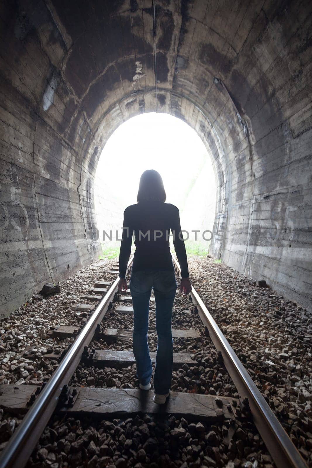 Man silhouetted in a tunnel standing in the center of the railway tracks looking towards the light at the end of the tunnel in a conceptual image