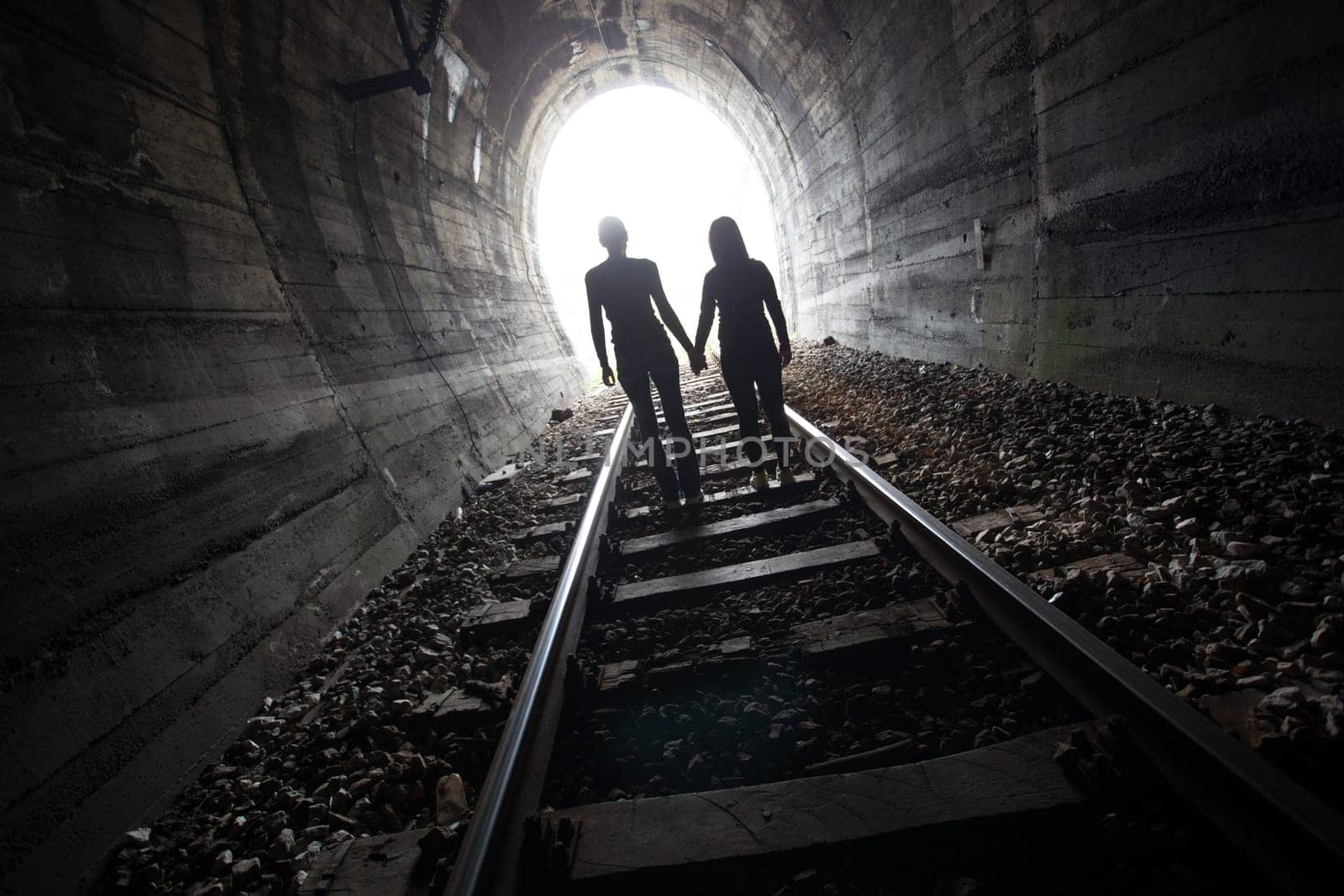 Couple walking hand in hand along the track through a railway tunnel towards the bright light at the other end, they appear as silhouettes against the light