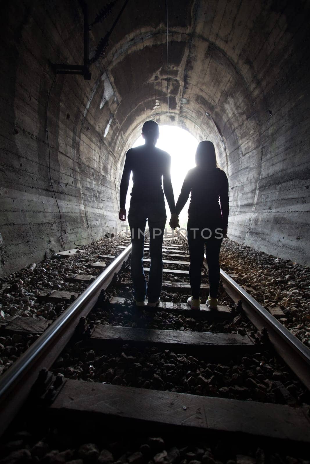 Couple walking hand in hand along the track through a railway tunnel towards the bright light at the other end, they appear as silhouettes against the light