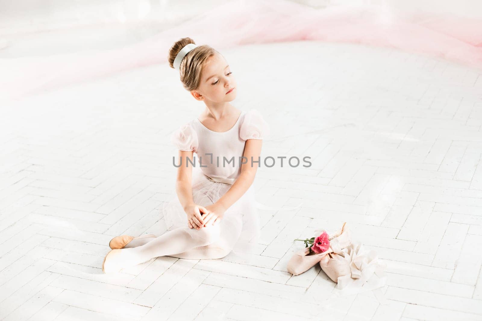 The little balerina in white tutu in class at the ballet school sitting on the wooden floor with flowers