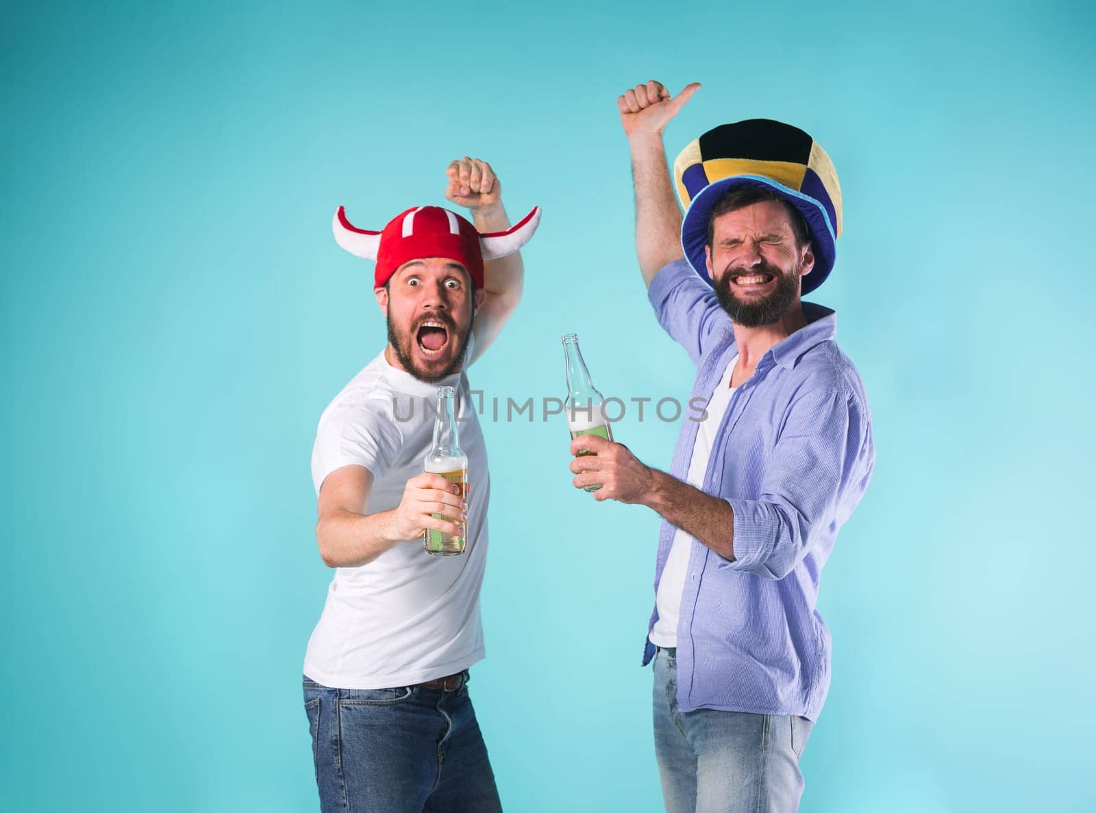 Two excited male friends celebrate watching sports on television with beer over blue studio