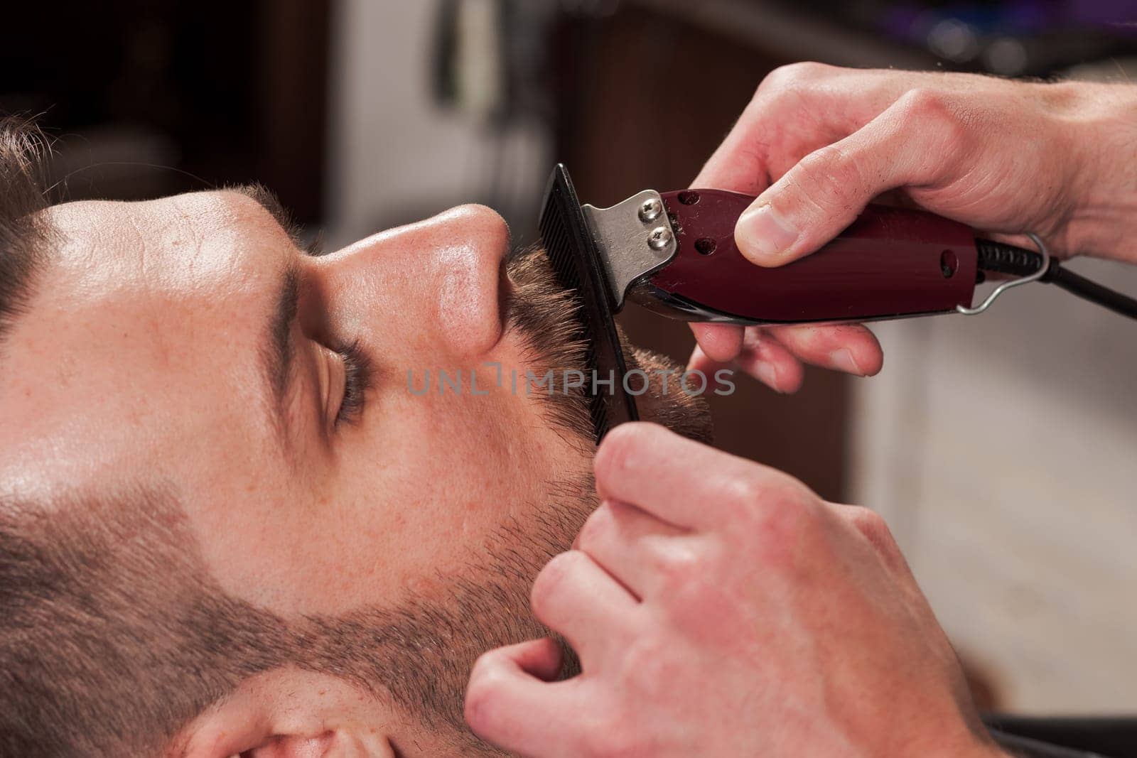 Hipster client visiting barber shop. The hands of young barber making the cut of beard