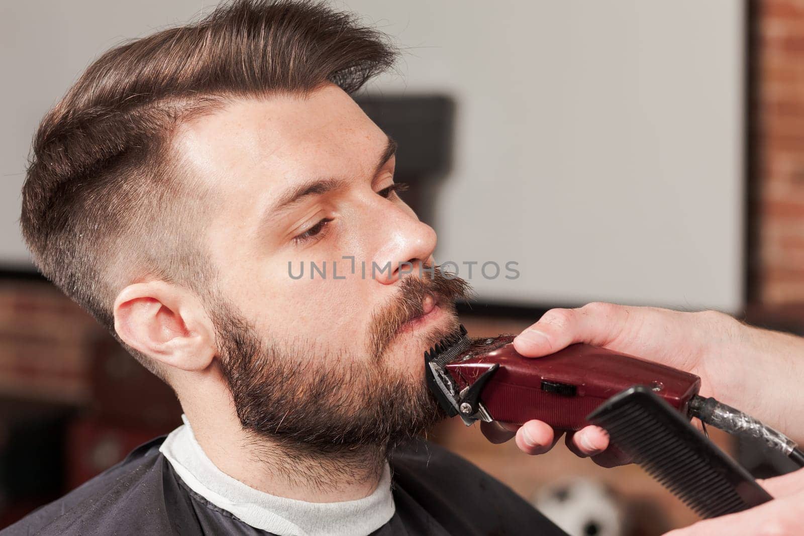 Hipster client visiting barber shop. The hands of young barber making the cut of beard